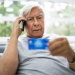 An older man holds up a credit car while speaking on a cellular telephone